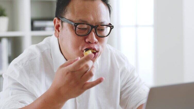 亚洲胖子在用笔记本电脑工作时吃甜甜圈、甜食、垃圾食品，不健康的饮食观念