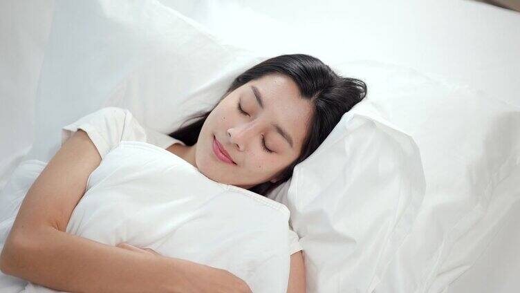 美丽的亚洲女人躺在床上。年轻的女人睡在舒适舒适的新鲜的床上柔软的枕头白色亚麻布。早上在卧室里。健康休息理念。