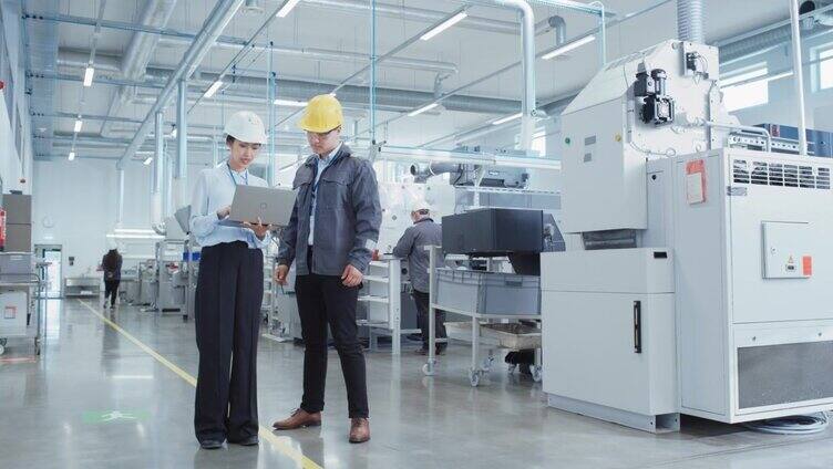 两个年轻的亚洲重工业工程师戴着安全帽站在一个工厂里，拿着笔记本电脑讨论工作过程。两名在生产设施工作的制造员工。