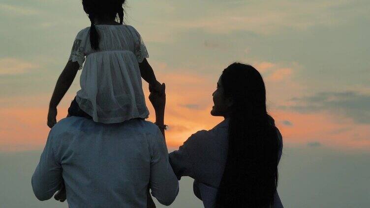 后视镜，一个4岁的亚洲女孩骑在父亲的脖子上，母亲牵着她的手，看着海滩上的日落。幸福的家庭假期。援助之手的概念。