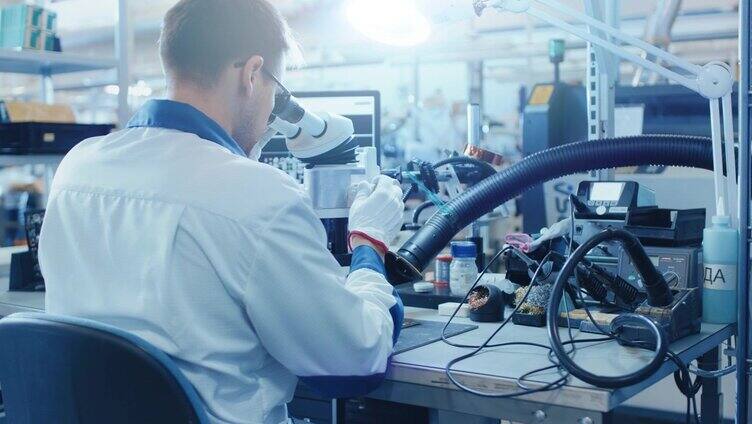 一名穿着白色工作服的电子厂工人通过数码显微镜检查一块印刷电路板。高科技工厂设施。