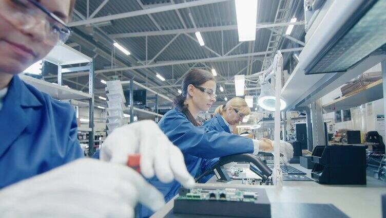穿着蓝色工作服和防护眼镜的电子厂女工正在用螺丝刀组装笔记本电脑的主板。拥有多名员工的高科技工厂设施。