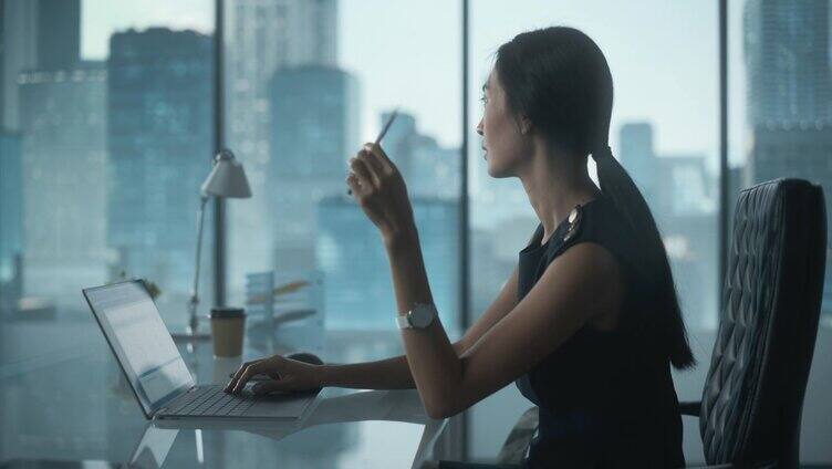 成功的女商人穿着时尚的裙子坐在办公桌在现代办公室，使用笔记本电脑，旁边的窗户与大城市的景色。成功的财务经理计划工作项目。