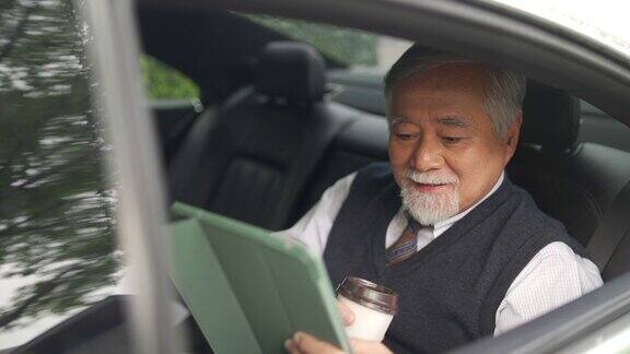 4K亚洲资深商人坐在汽车后座一边喝着热咖啡一边在数码平板电脑上工作