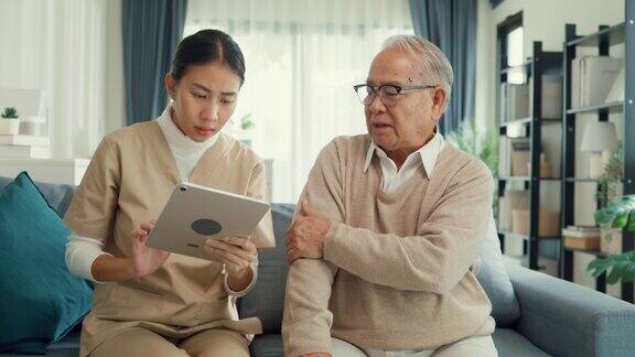 年轻的亚洲女性看护者手持平板电脑照顾平静的生病的老年男性患者在家里的客厅