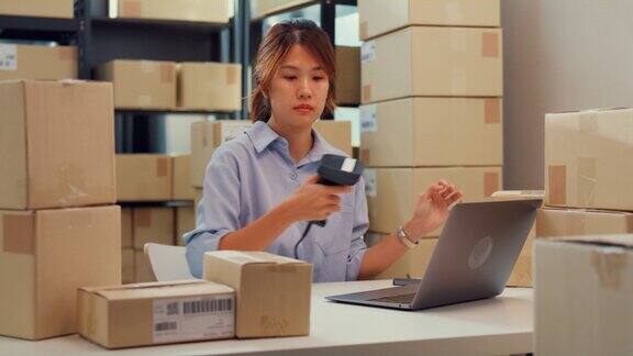 亚洲女商人坐在货架前使用条形码扫描纸盒上的客户数据登记在线代码在笔记本电脑上订购明细以便在仓库快递