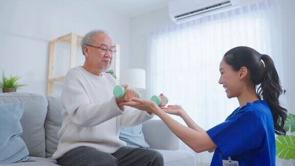 亚洲年长男子在护士的支持下做物理治疗老年男性患者坐在客厅的沙发上使用哑铃锻炼与治疗师医生在养老院照顾