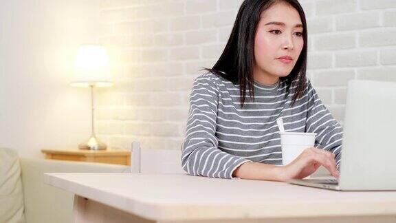 亚洲自由职业商业女性吃方便面而工作在笔记本电脑在客厅在家庭办公室晚上年轻的亚洲女孩坐在办公桌上加班享受放松的时光女性工作