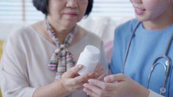 在家访护理服务中年轻的护理人员穿着手术服向年长的亚洲妇女展示药瓶亚洲老年人与辅助生活药物监测坐在家里交谈