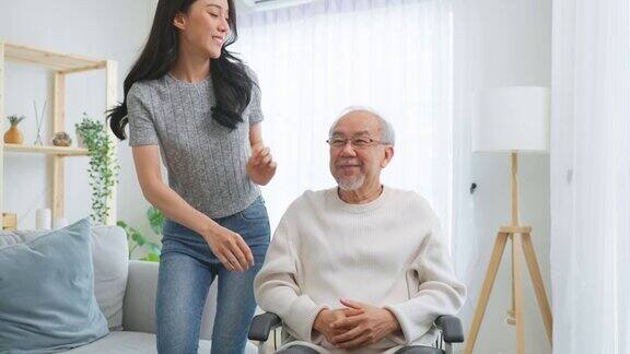 亚洲年轻的女儿坐在轮椅上照顾年长的男性美丽的女孩帮助和支持老人、老、成熟的爷爷病人在客厅做理疗健康的房子