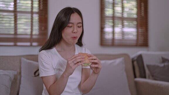 年轻饥饿的亚洲妇女吃着垃圾食品汉堡包在家里吃着快餐展示着减肥食品的选择