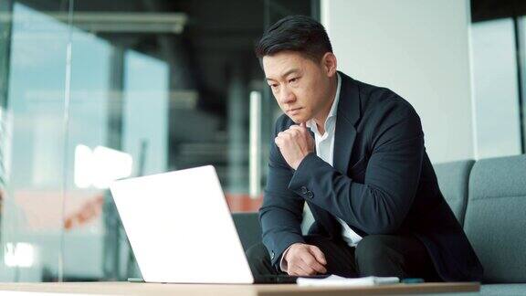 照片中有思想的亚洲商人在现代办公桌上用笔记本电脑工作自信、专注、沉思的男士在室内穿着正装用灵感解决问题