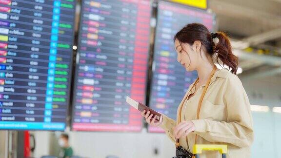 年轻的亚洲女子拿着护照和登机牌在国际机场看航班信息板检查她的航班