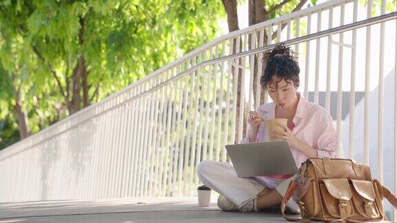 一位女商人一边吃面条盒饭一边用笔记本电脑在公园里工作