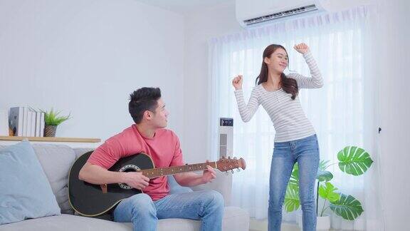 亚洲年轻情侣在家里的客厅里一起弹吉他迷人浪漫的新婚男女坐在沙发上玩乐器在家里一起
