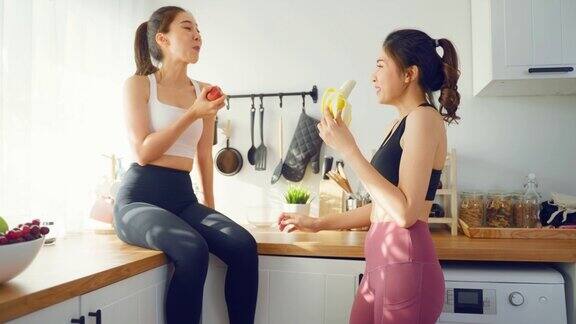 亚洲活跃的两个女人姐妹穿着运动服在厨房吃苹果年轻美丽的女孩妹妹心情愉快享受吃水果健康的食物来饮食和减肥的保健在家里