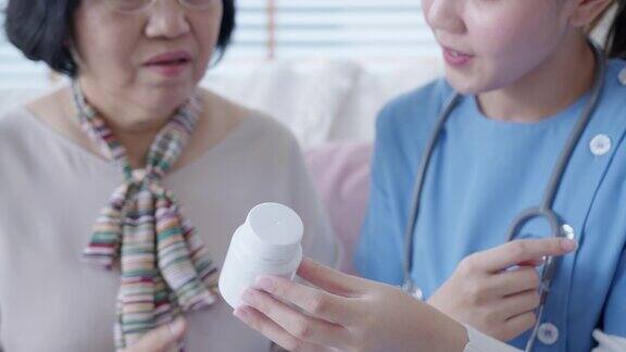 在家访护理服务中年轻的护理人员穿着手术服向年长的亚洲妇女展示药瓶亚洲老年人与辅助生活药物监测坐在家里交谈
