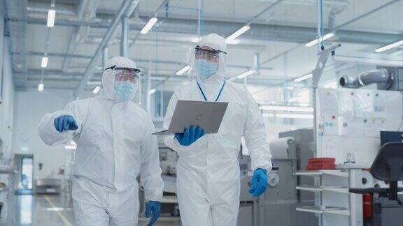 两名科学家穿着无菌工作服和口罩走在重工业工厂使用笔记本电脑检查工业机器设置和配置生产功能
