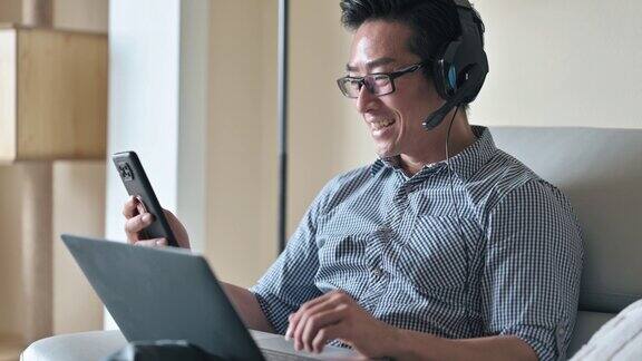 亚洲华人男性在家工作坐在沙发上戴着耳机用智能手机聊天