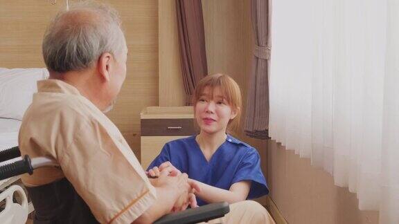 在医院照顾坐在轮椅上的成熟男性病人的亚洲护士年轻女子推着轮椅然后坐下来微笑着和老人交谈待客医疗服务理念