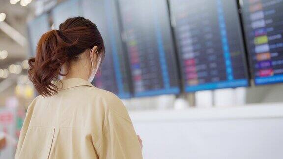 年轻的亚洲女子戴着口罩拿着护照和登机牌在国际机场看着航班信息板检查她的航班