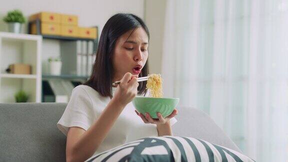 年轻的亚洲女人坐在沙发上拿着电视遥控器看电影吃着方便面在家放松的一天