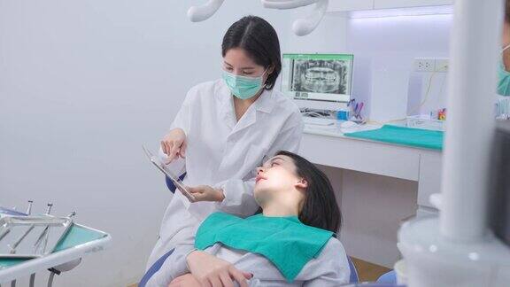 亚洲年轻女牙医拿着数码片给高加索年轻女孩病人建议或咨询医生讲解牙科诊所提供的口腔护理服务