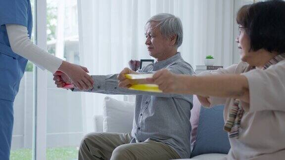 年轻的老年亚洲公民夫妇坐在沙发沙发上配合居家护理辅助生活搓洗护士使用阻力带练习为老年患者在家中进行理疗治疗