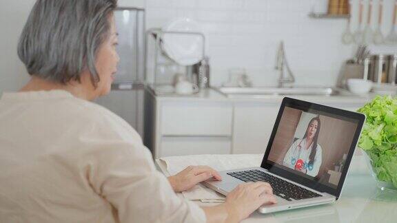 亚洲资深女性视频电话与医生在客厅在家老年病人咨询全科医生应用电脑医生和顾问在线和远程医疗