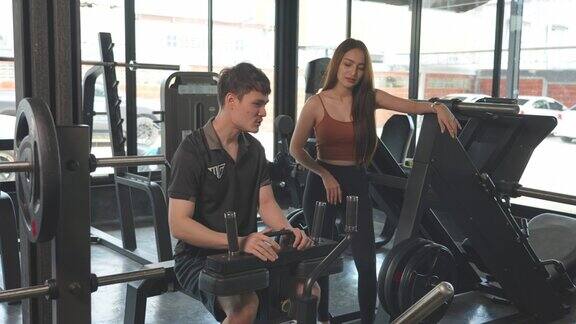 亚洲健身教练男性支持教和激励年轻美丽的初学者女运动员锻炼举重机活跃的健美女孩在体育馆体育馆锻炼身体为健康锻炼肌肉