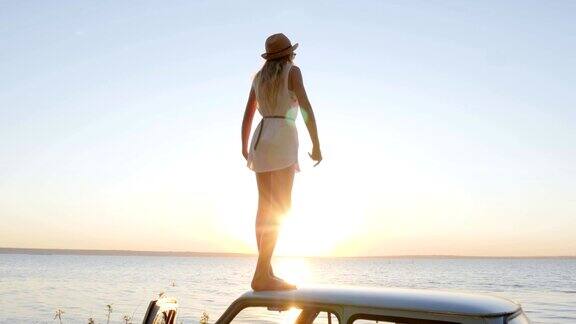 快乐的旅行女孩站在车顶双手张开欣赏日落自由的快乐在海边年轻女子站在举起的手