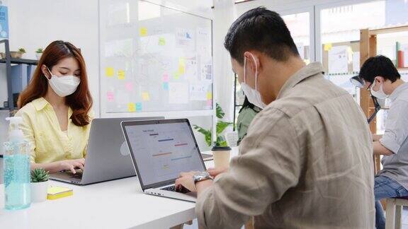 亚洲企业家在新常态下戴着医用口罩进行社交距离以预防病毒在办公室使用笔记本电脑工作