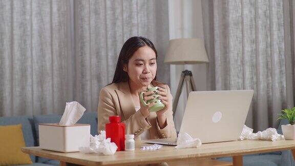 生病的亚洲女商人在家里的客厅用笔记本电脑工作时喝热水