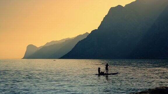 游客在平静的加尔达湖冲浪板上在日落的橙色天空下沿着山脉
