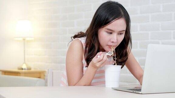 亚洲自由职业商业女性吃方便面而工作在笔记本电脑在客厅在家庭办公室晚上年轻的亚洲女孩坐在办公桌上加班享受放松的时光女性工作