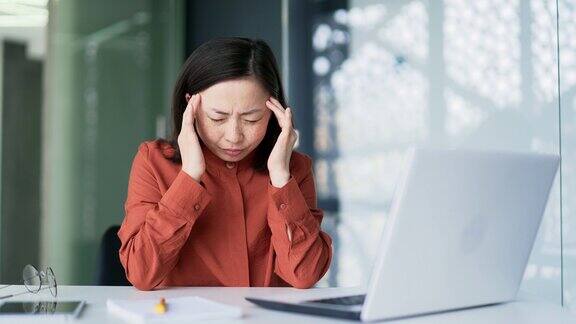 悲伤的亚洲女商人坐在办公室里用笔记本电脑工作时头痛