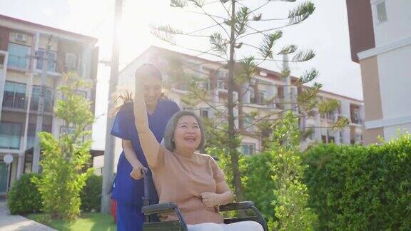 亚洲快乐的老病人奶奶在绿色公园户外玩护理员微笑着推着坐在轮椅上举起手臂的老人享受着养老院的炉边护理活动