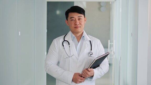 中国医生的特写肖像在宽敞明亮的医院走廊拿着文件夹文件在办公室走廊里身穿白色白大褂的成功医生看着镜头微笑