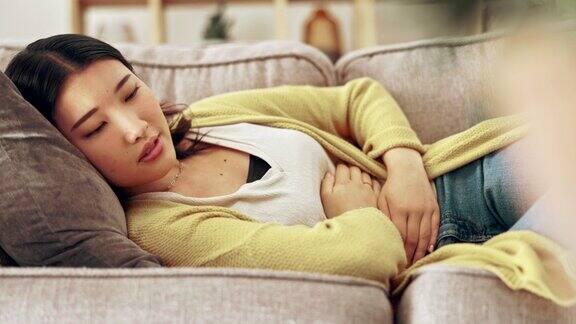疼痛经期和亚洲妇女用手放在肚子上怀孕月经或腹部痉挛健康消化问题和女孩在沙发上抱着腹部子宫内膜异位症经前综合症和疼痛在家里