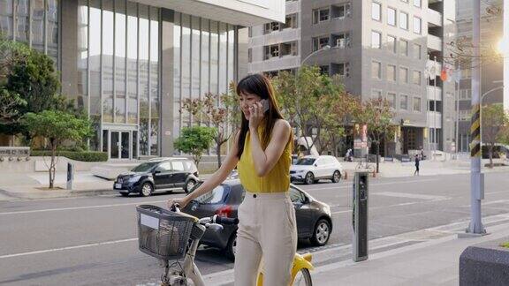 一名亚洲女子推着一辆共享单车在城市街道上用智能手机与同事或朋友交流