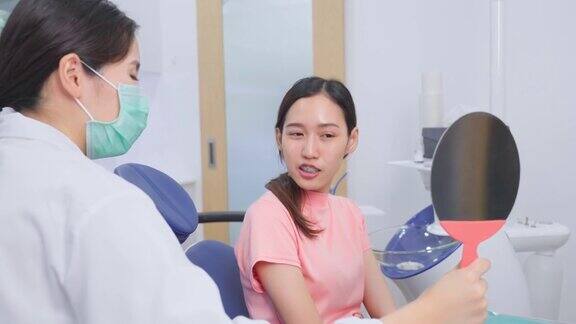 亚洲牙齿矫正医师扶着镜子让年轻的病人在调整线后检查她的牙套女人看着镜子张开嘴看着她的牙齿牙科诊所的口腔护理