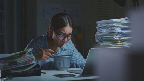 年轻的亚洲办公室职员在办公室用笔记本电脑边吃泡面边工作晚上加班