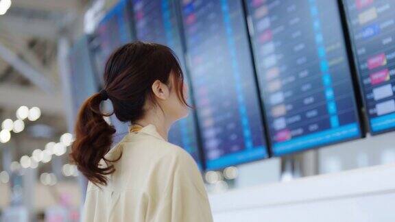 年轻的亚洲女子手持护照和登机牌在国际机场查看航班信息板查询她的航班