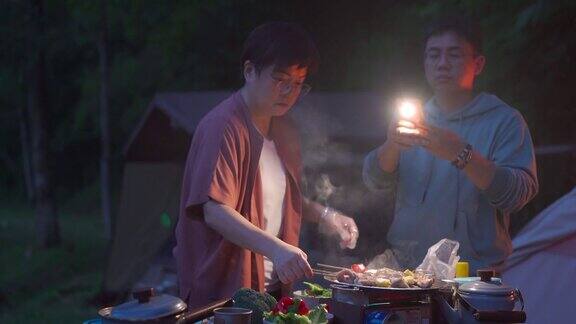 一个亚洲中国女人在晚上露营时准备烧烤而她的丈夫用他的智能手机点亮了场景