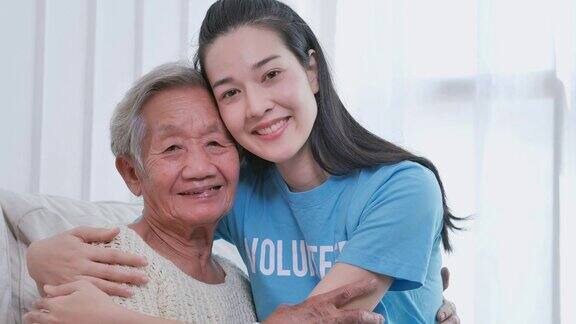 描绘亚洲年轻女性志愿者拥抱亚洲年长女性微笑着看着家里的相机志愿服务