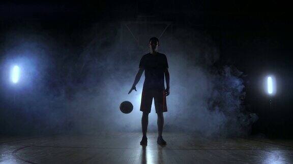 篮球运动员穿着运动服红色短裤和蓝色t恤走在一个黑暗的篮球场上背光从烟雾中出来敲打一个篮球在地板上看着慢镜头