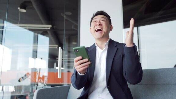 高兴高兴的亚洲商人穿着西装庆祝手机获胜兴奋万分的幸运高管获胜者在短信中收到好消息使用智能手机