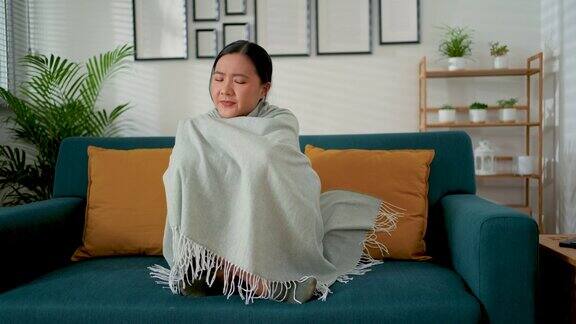 亚洲女人觉得冷裹着毯子坐在家里客厅的沙发上