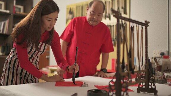 一位亚洲华人男子练习书法为即将到来的中国新年庆祝家庭装饰目的繁荣和良好的措辞写在一张红色的纸上并教他的女儿