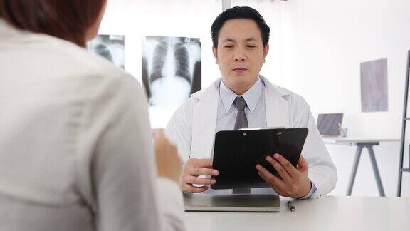 严肃的亚洲男医生正在用剪贴板与女病人坐在医院办公室的桌子前传递好消息谈论结果或症状